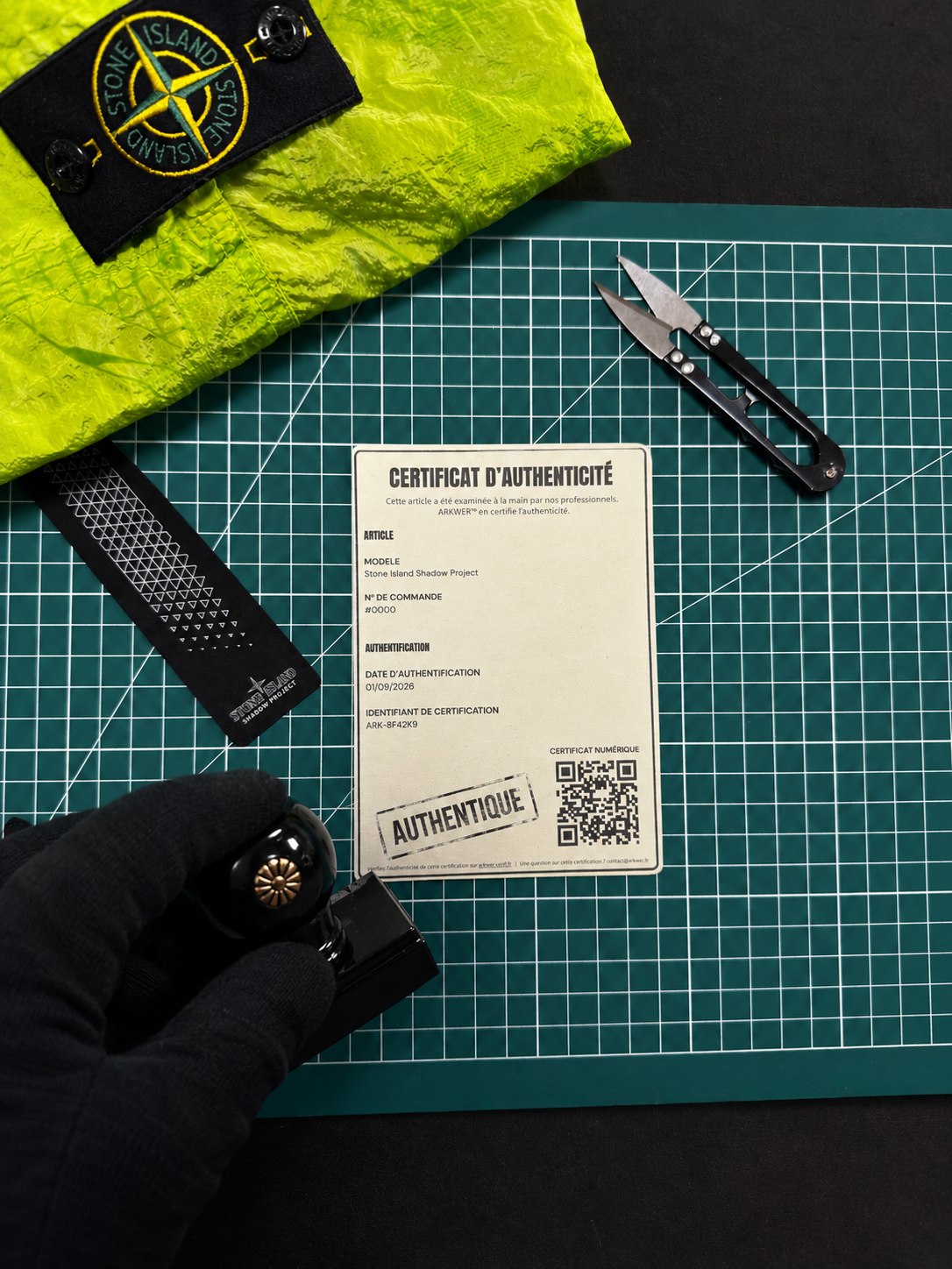 Certificat d'authenticité ARKWER posé sur un tapis de découpe vert, à côté d'une pièce Stone Island Shadow Project ; une main gantée tient le tampon.