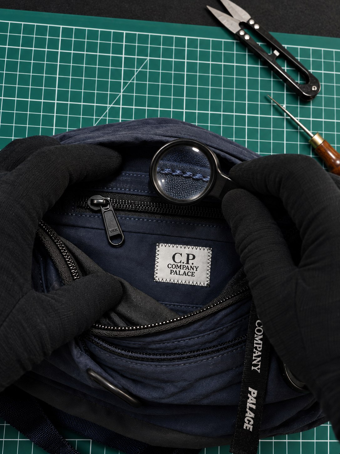 Mains gantées ouvrant une sacoche C.P. Company Palace ; une loupe d'horloger grossit les surpiqûres bleu marine, sur un tapis de découpe vert.