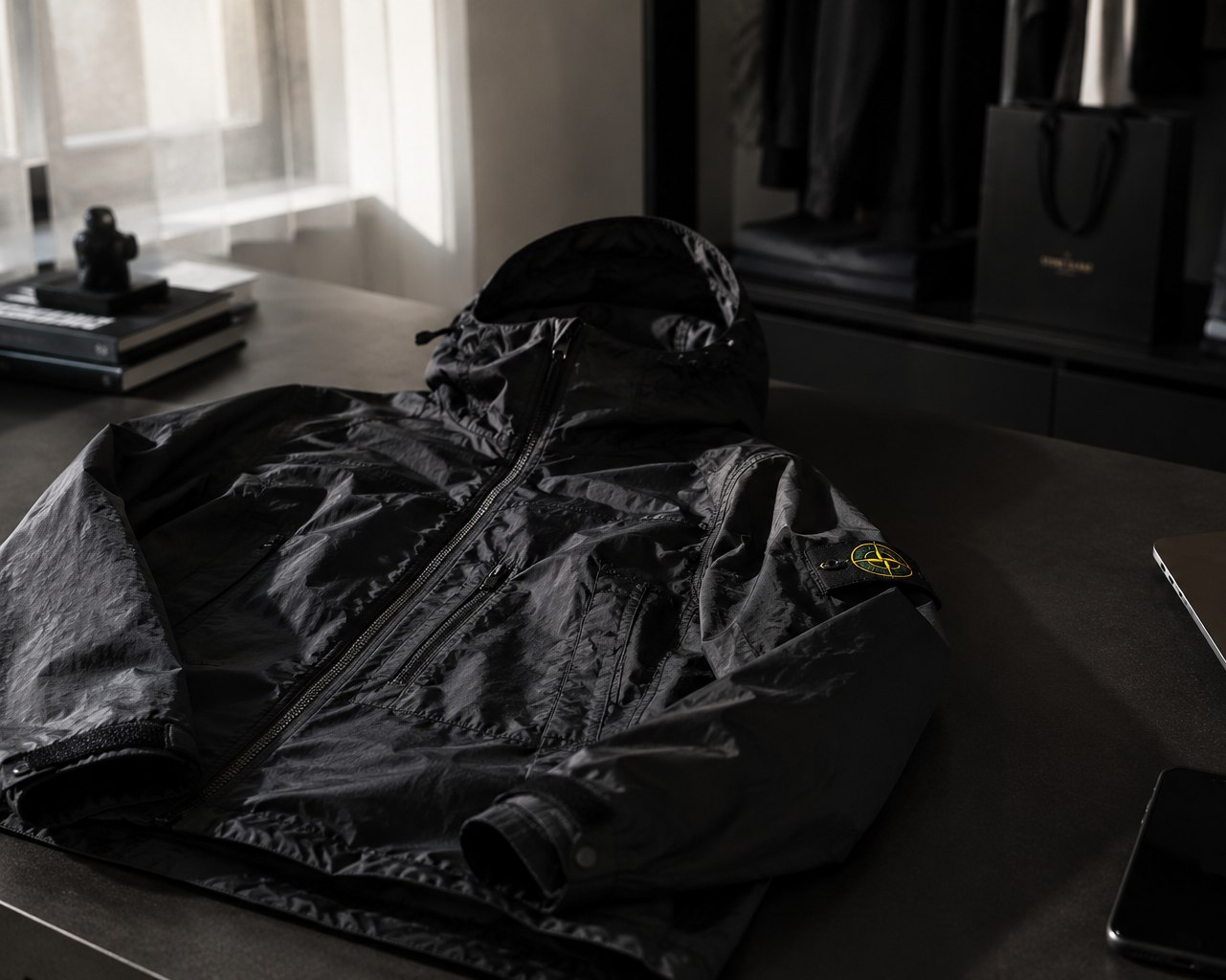 Veste à capuche Stone Island, badge jaune sur la manche, posée sur un plan de travail sombre devant une fenêtre voilée.